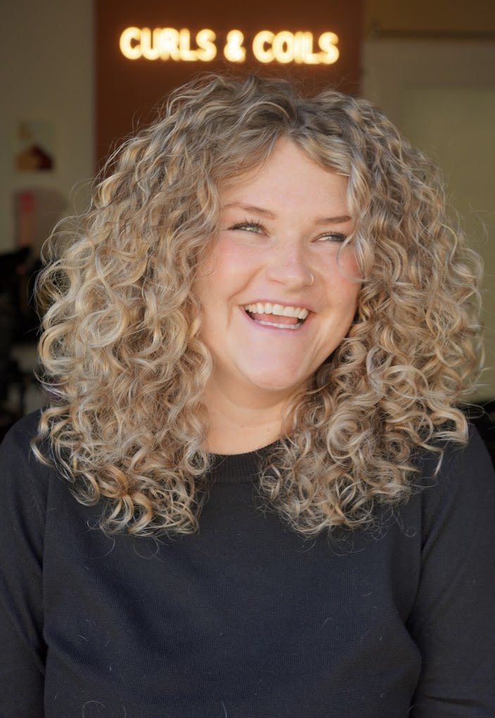 A smiling guest with long, defined blonde curls in front of the glowing Curls & Coils sign