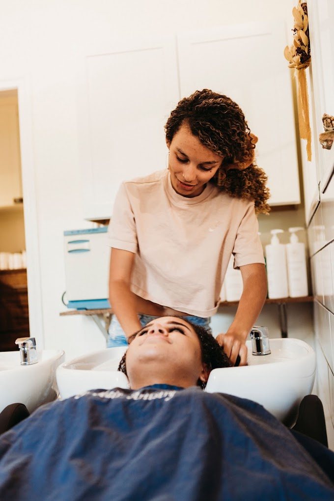 A curly-haired stylist washing a guest’s hair at the shampoo bowl